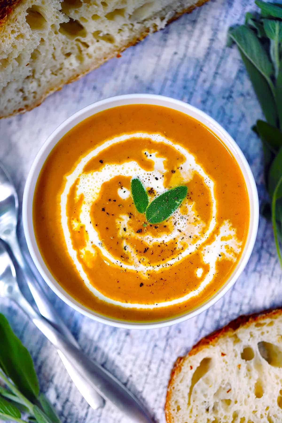 Overhead photo of a bowl of pumpkin soup drizzled with cream and garnished with fresh sage leaves, with bread around it.