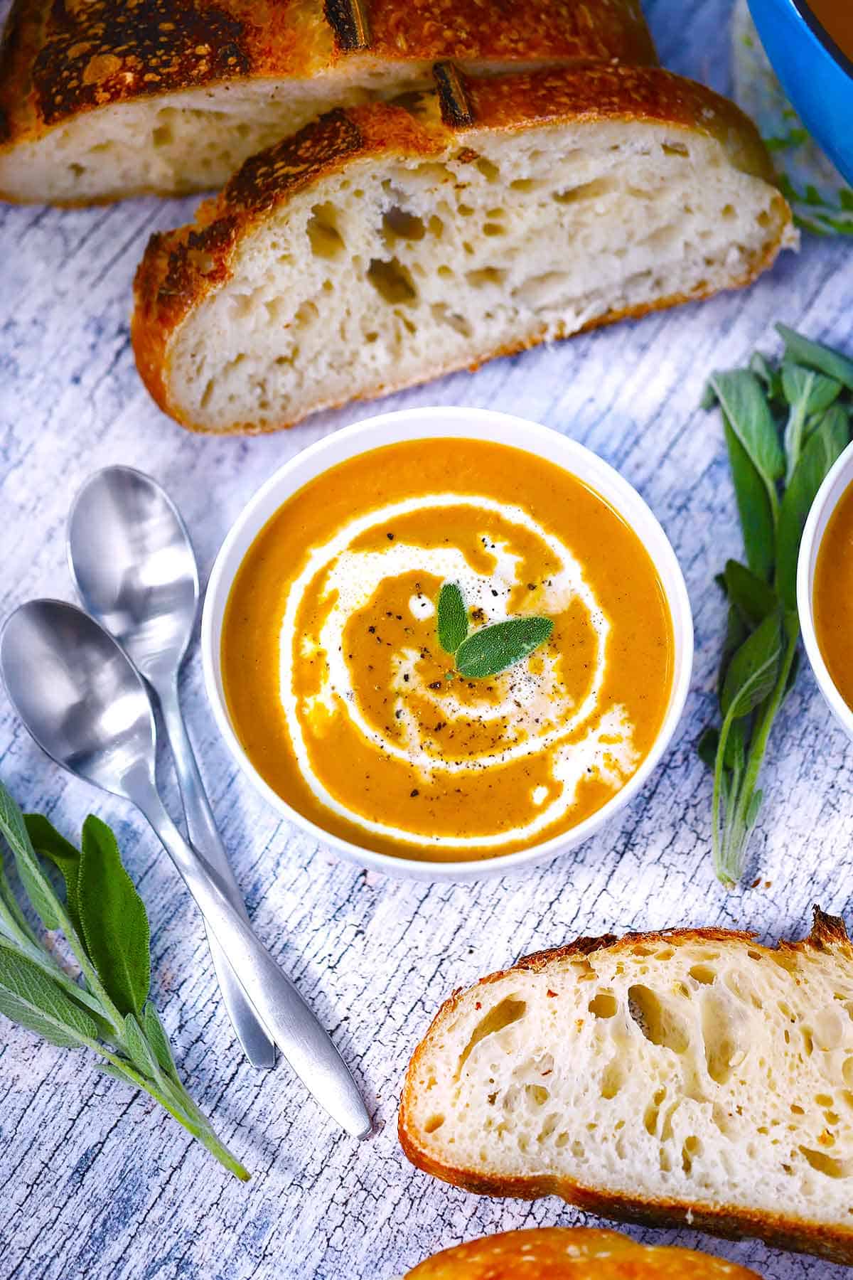 A white bowl with pumpkin soup and sourdough bread and fresh sage scattered around it.