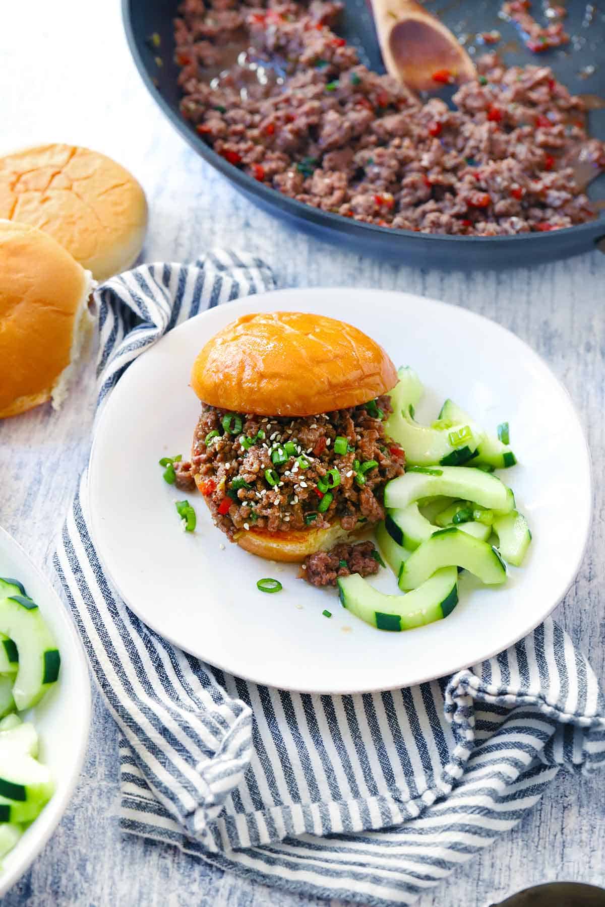 A korean bbq sloppy joe sandwich on a white plate with a skillet in the background,