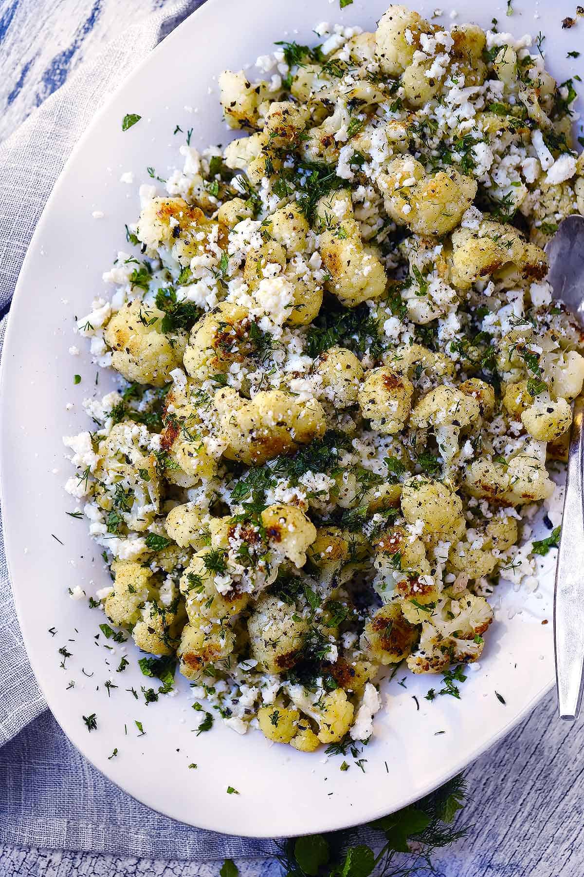Overhead photo of a platter of Greek Style Roasted Califlower topped with Feta Cheese, Herbs, and Lemon.
