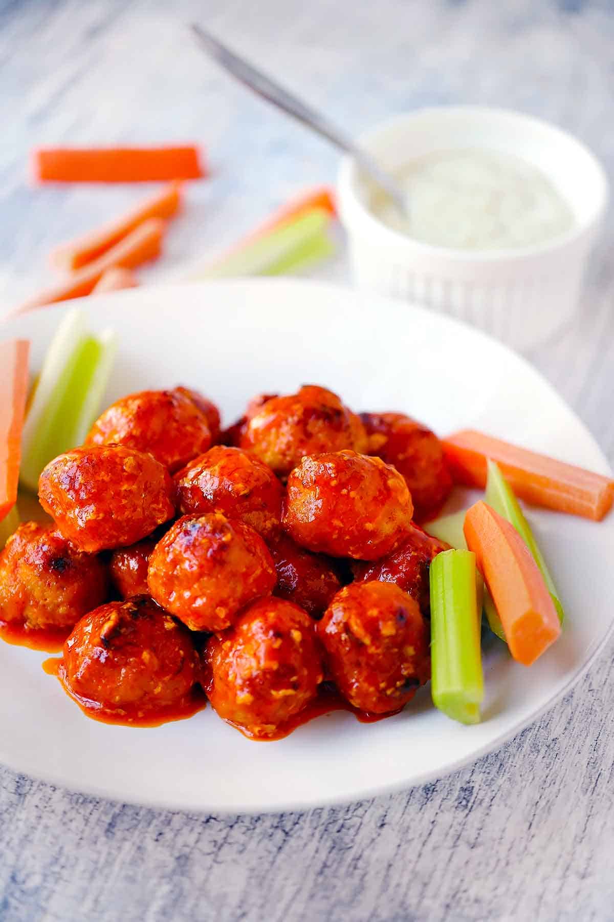 A white plate with buffalo chicken meatballs, celery, and carrots with blue cheese dressing in background.