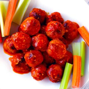 Square photo of buffalo chicken meatballs with celery and carrots on a white plate.