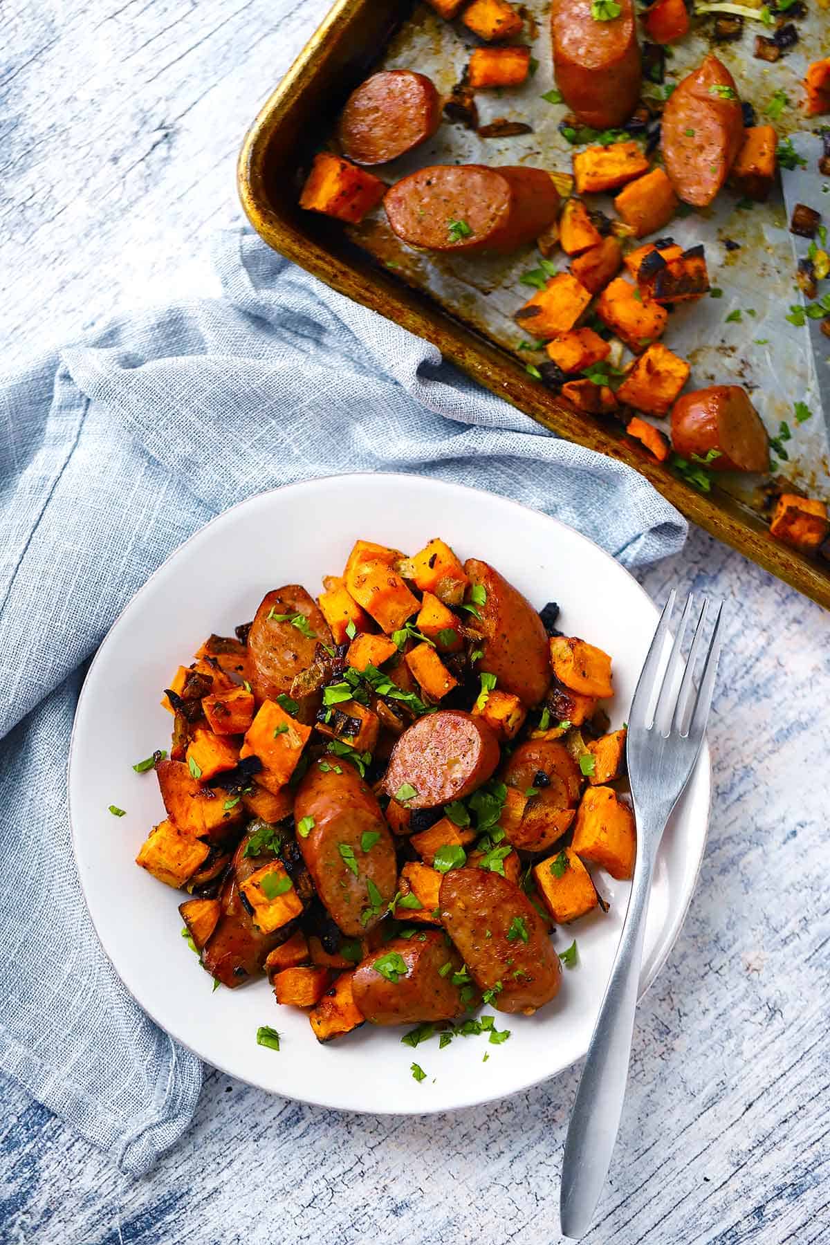 Sausage and sweet potatoes on a white plate next to a sheet pan.