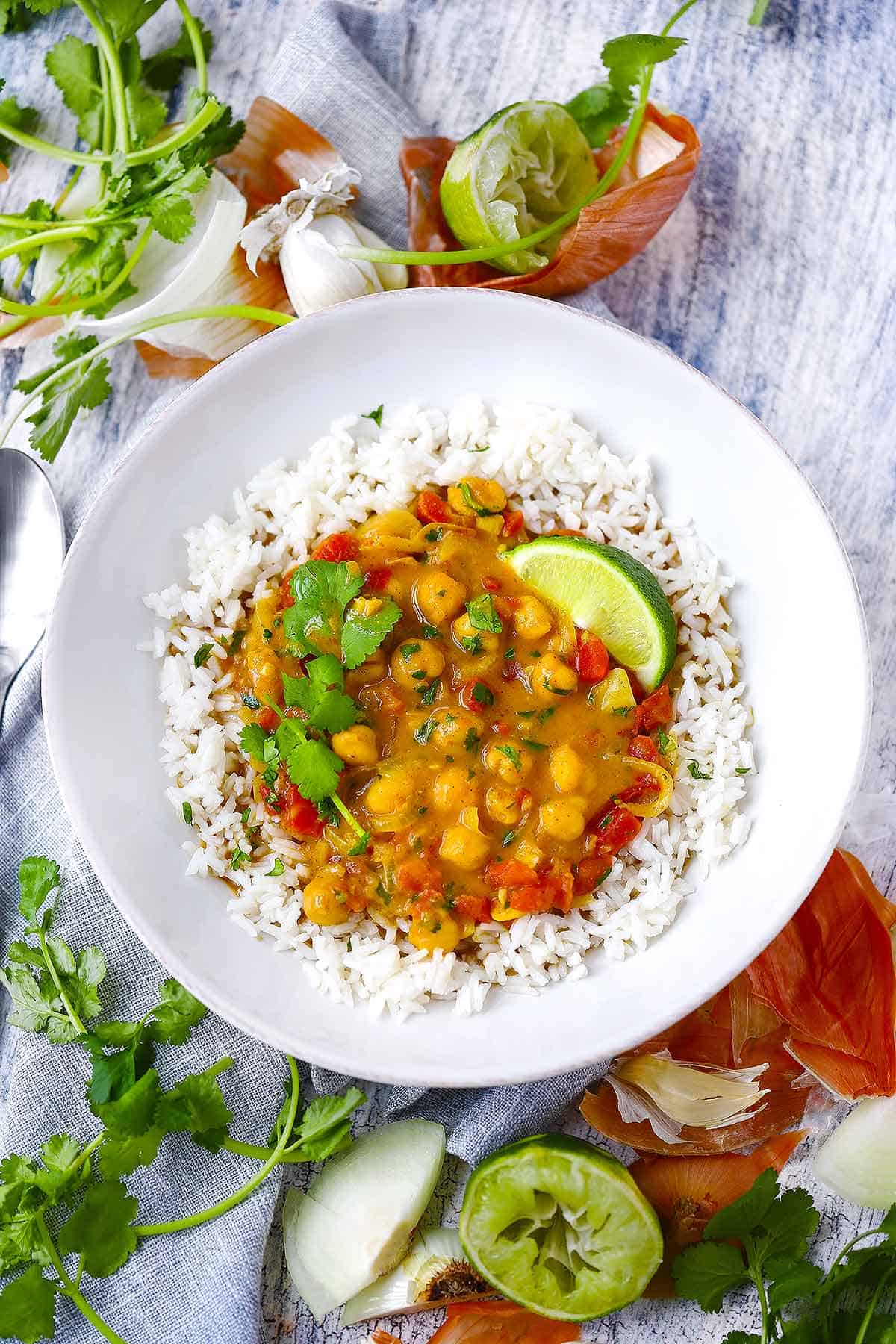 Overhead photo of chickpea curry over rice in a white bowl with lime and cilantro and ingredients scattered around.