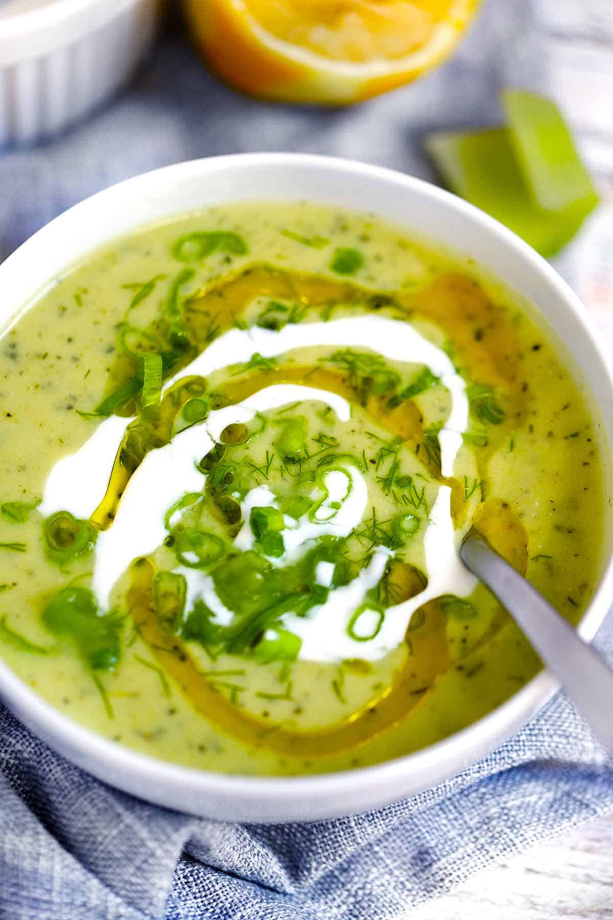 A white bowl with zucchini soup and a spoon in it with olive oil, yogurt, and herbs on top.