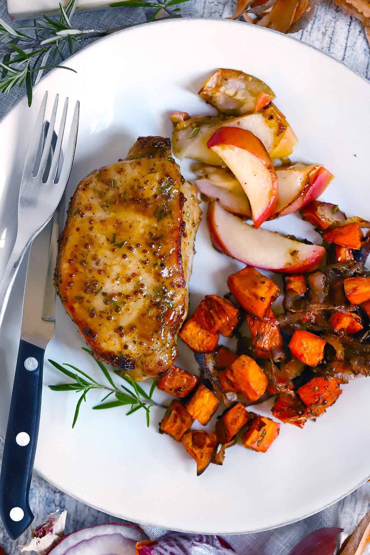 A plate with a pork chop, roasted apples, and sweet potatoes and a fork and knife on the side.