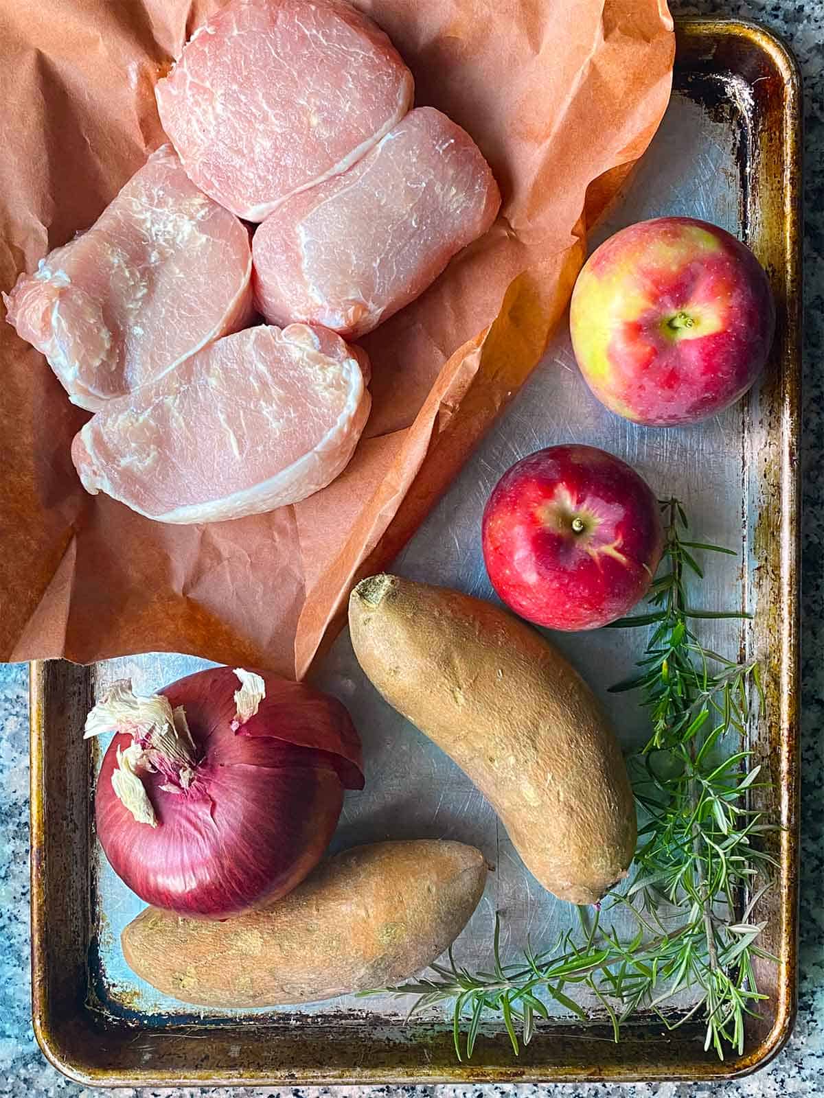 Boneless pork chops, apples, sweet potatoes, and an onion, with a sprig of rosemary, to show ingredients for sheet pan pork chops.