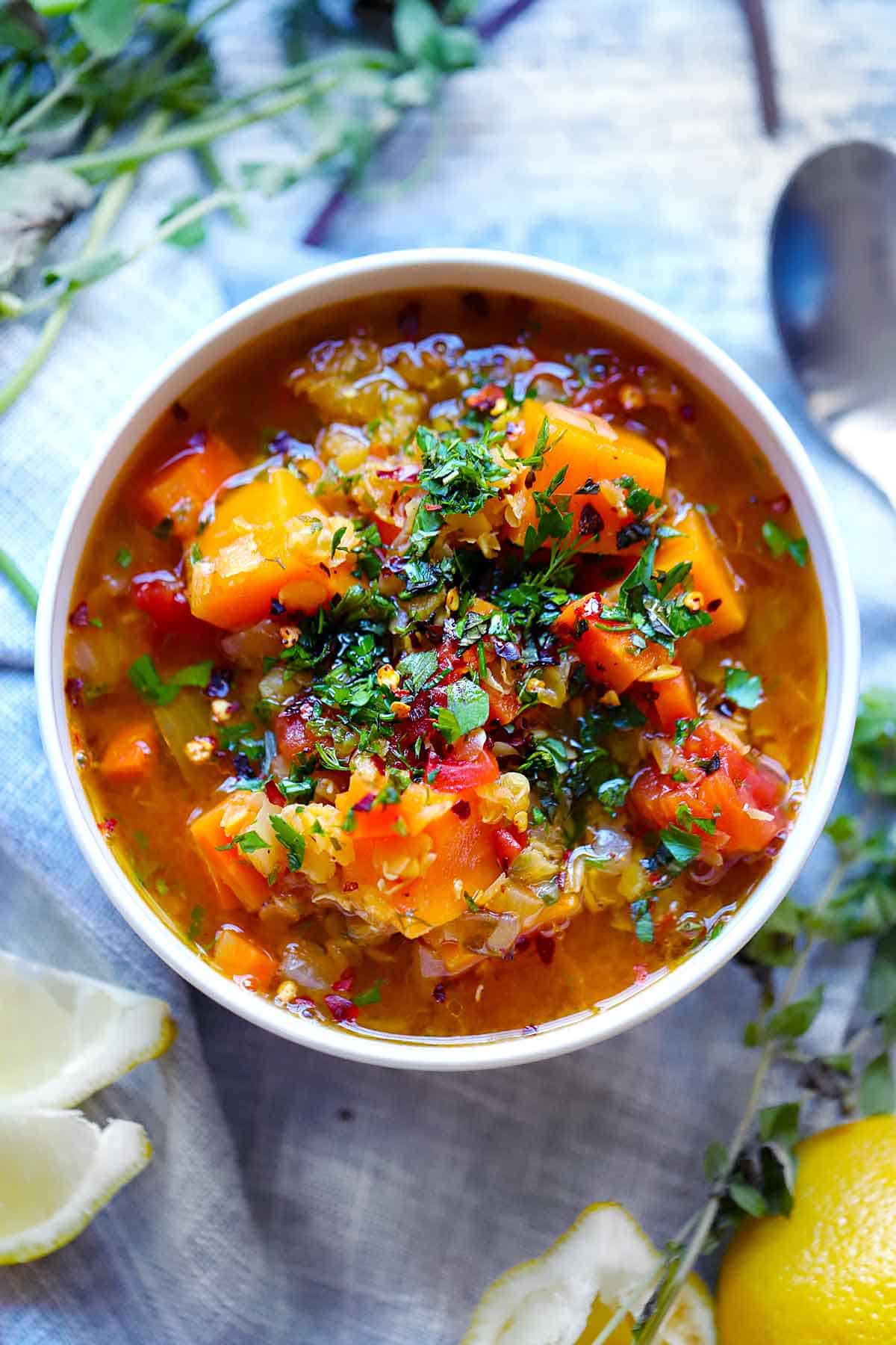 Overhead photo of a white bowl of red lentil soup with sweet potatoes, topped with herbs, lemon wedges surrounding it.