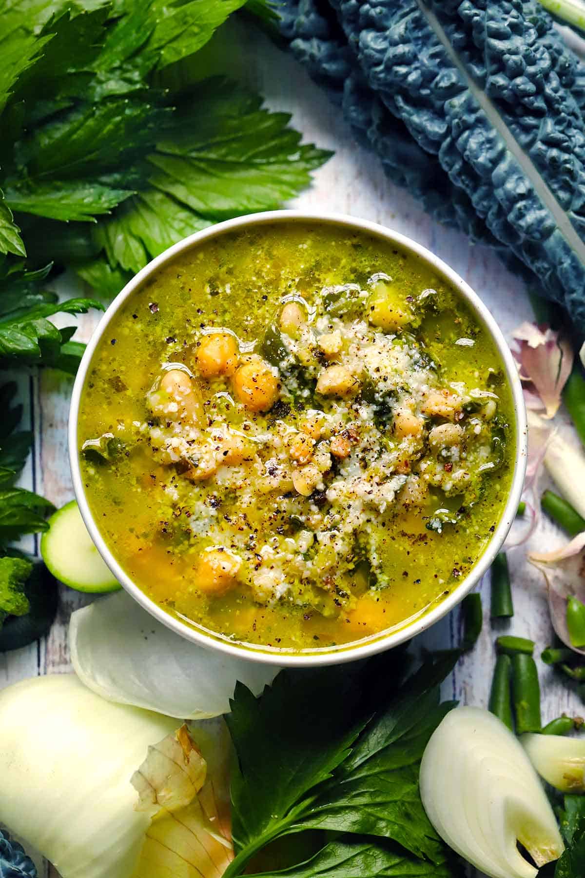 Overhead view of green pesto minestrone soup with chickpeas, white beans, and grated Parmesan in a white bowl, surrounded by fresh parsley, kale, and zucchini.