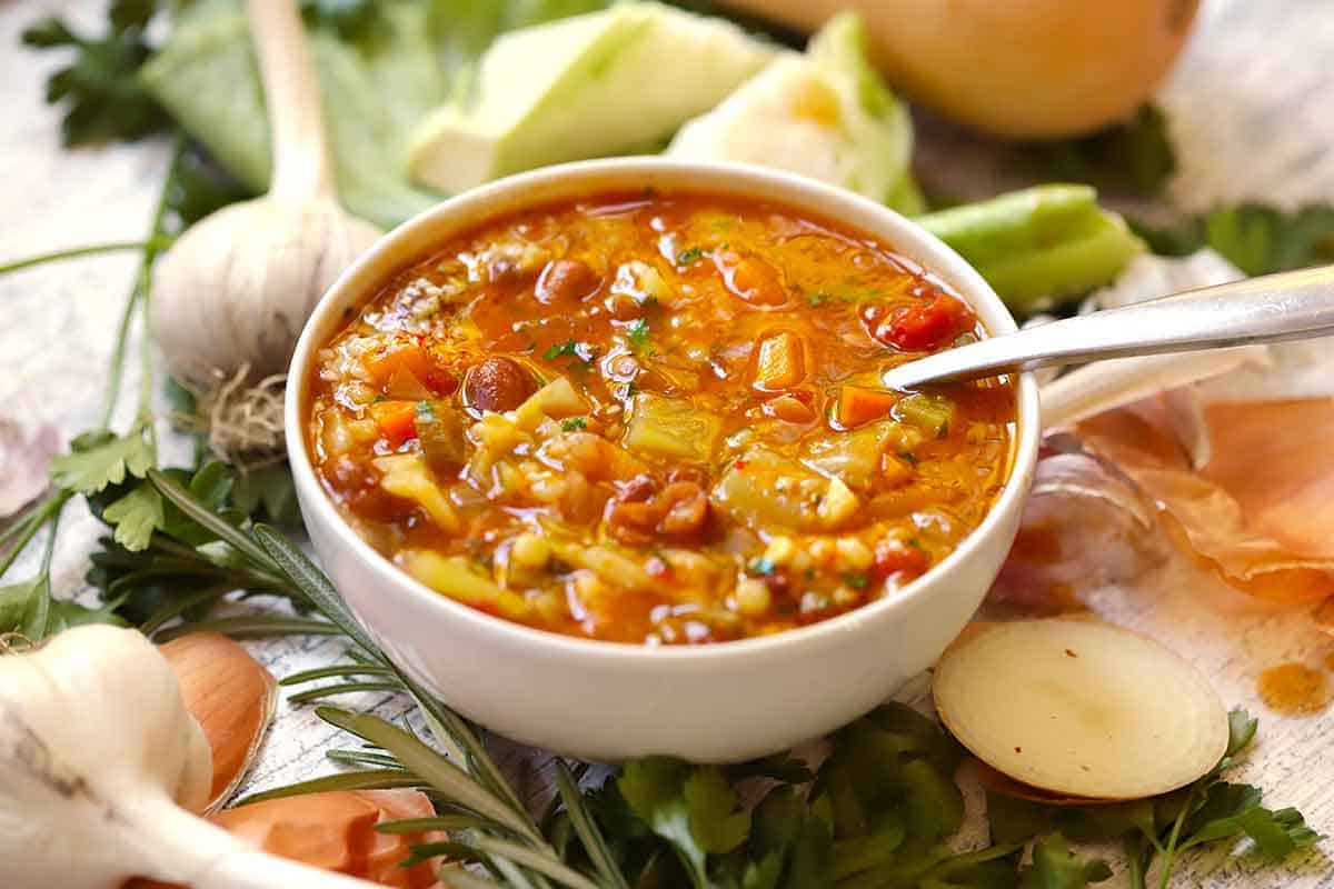 Angled view of a white bowl of autumn minestrone with squash, beans, cabbage, and barley in tomato broth, with a spoon, garlic, rosemary, and parsley nearby.