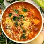 Square overhead photo of autumn harvest minestrone soup with squash, beans, grated cheese, red pepper flakes, and parsley in a white bowl.