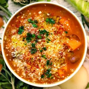 Square overhead photo of autumn harvest minestrone soup with squash, beans, grated cheese, red pepper flakes, and parsley in a white bowl.