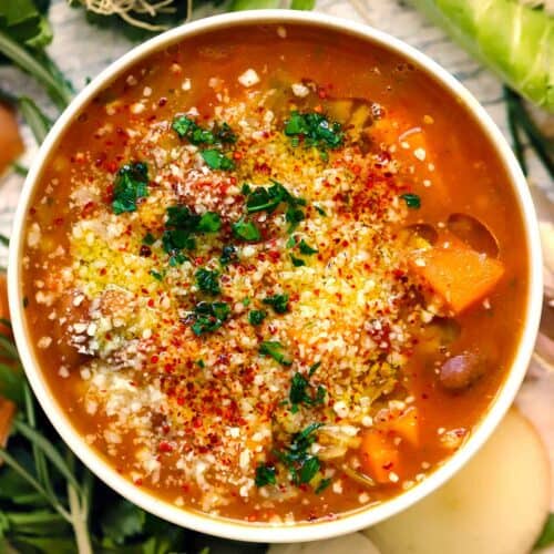 Square overhead photo of autumn harvest minestrone soup with squash, beans, grated cheese, red pepper flakes, and parsley in a white bowl.