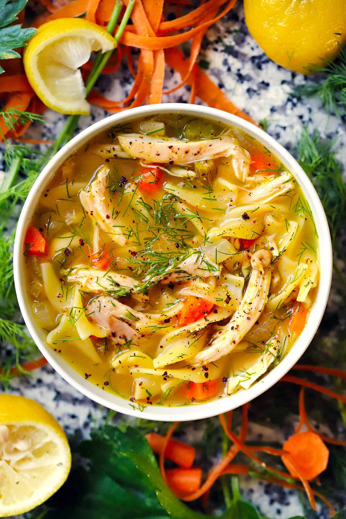 Overhead view of chicken noodle soup with shredded chicken, carrots, noodles, and fresh dill in a white bowl, surrounded by lemon wedges and carrot ribbons.