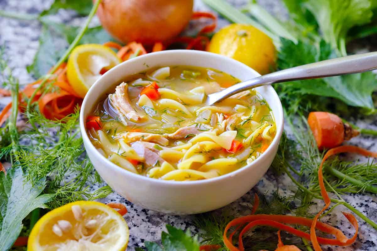 Angled view of a bowl of chicken noodle soup with a spoon, surrounded by lemons, carrot ribbons, and fresh herbs.