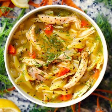Square overhead photo of chicken noodle soup with shredded chicken, carrots, noodles, and dill in a white bowl.