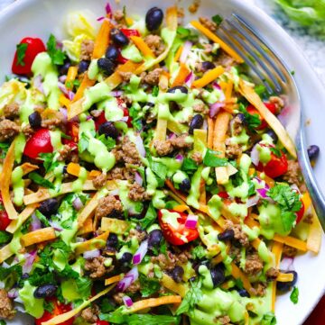 Square photo of an overhead view of a bowl of taco salad with avocado cilantro dressing, ground turkey, black beans, and tortilla strips.
