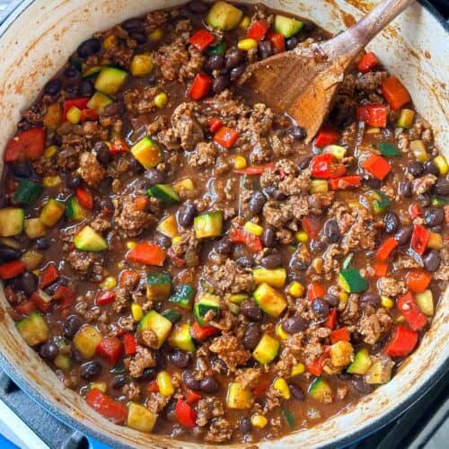 Overhead square photo of a pot of turkey chili with veggies and black beans with a wooden spoon.