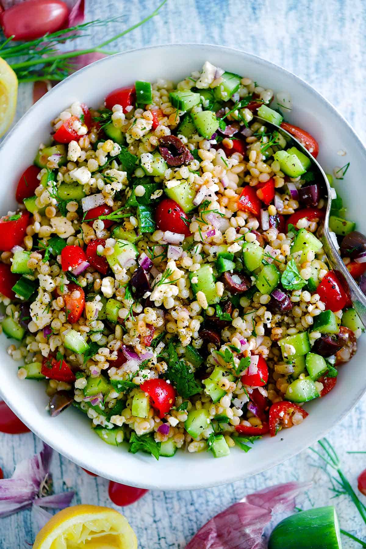 Overhead photo of a bowl of Israeli couscous salad with Mediterranean ingredients like cucumber, tomato, and feta, in a white bowl.