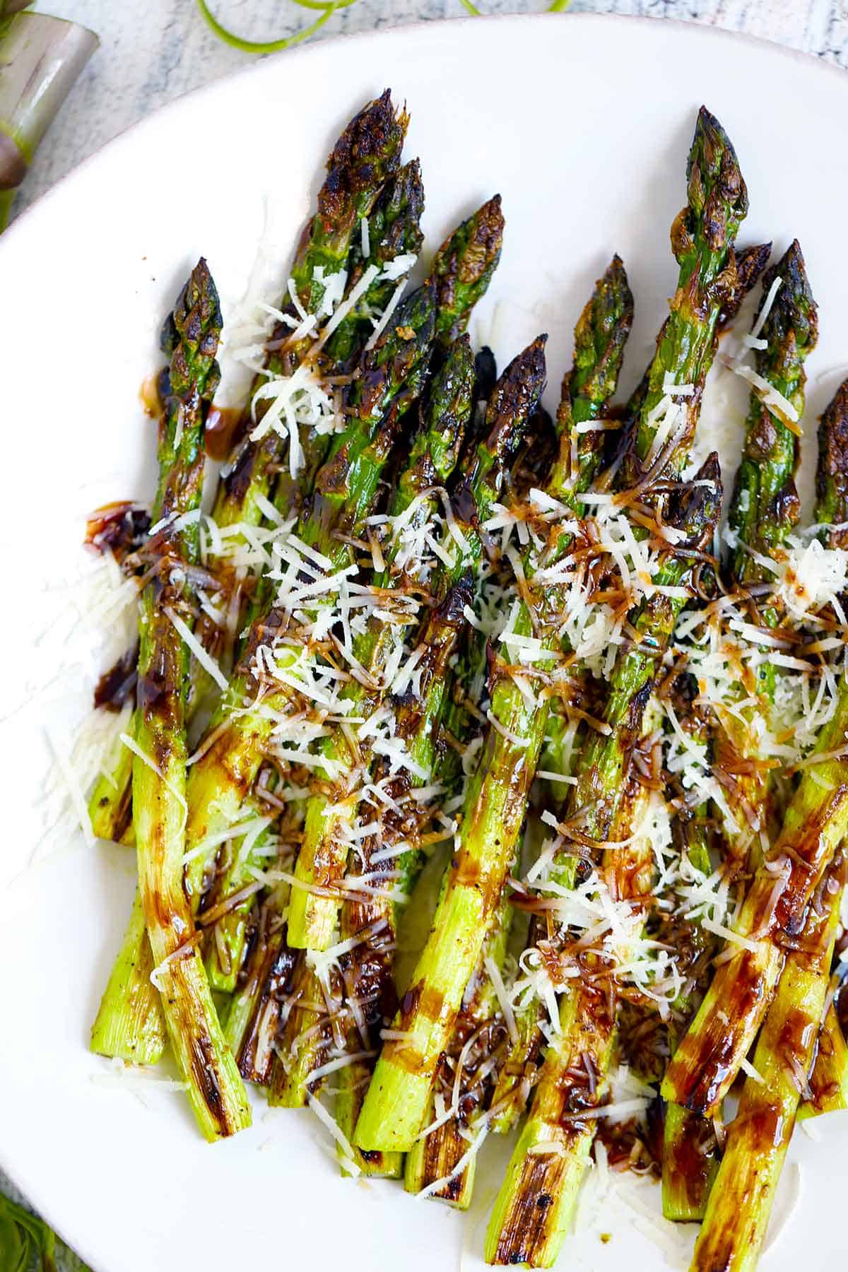 Overhead photo of roasted asparagus on a white plate topped with parmesan cheese and balsamic glaze.