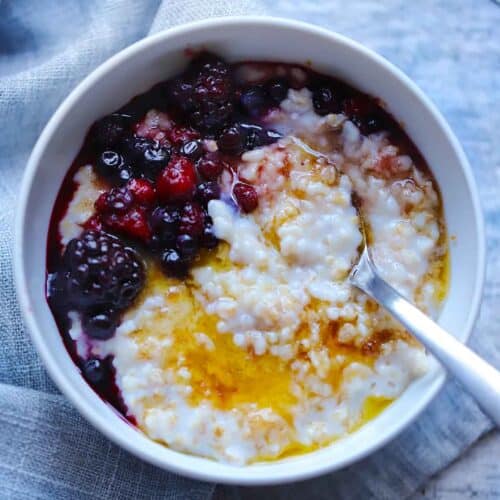 Square photo of a bowl of steel cut oatmeal with berries, brown sugar, and butter on top and a spoon in the bowl.