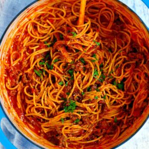 Square photo of an overhead view of a pot with spaghetti and meat sauce garnished with parsley in a blue Dutch oven.