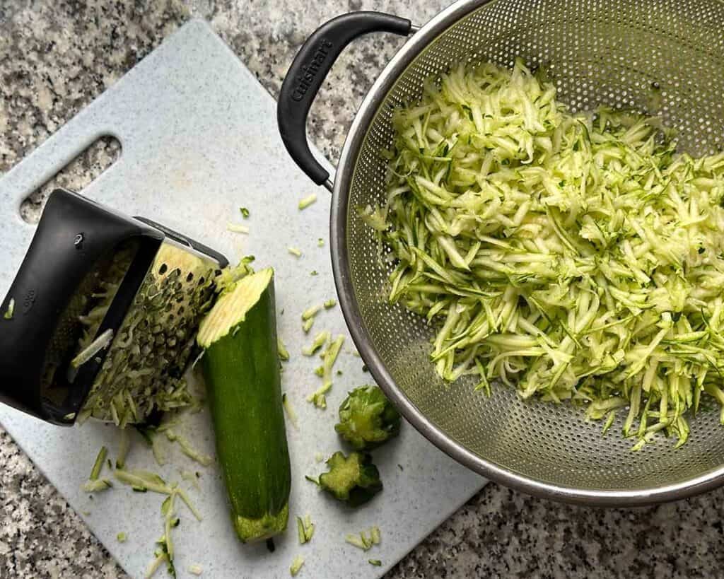 A colander over a bowl filled with grated zucchini, with a box grater and zucchini next to it.