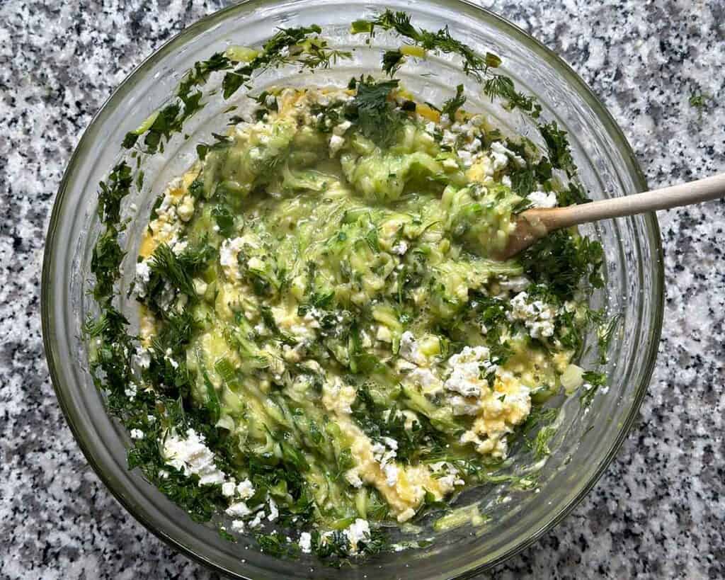 Mixing eggs, zucchini, herbs, green onions, milk, olive oil, and feta cheese together in a large bowl.