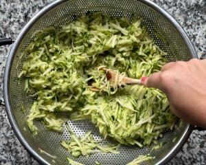 Squeezing liquid out of grated zucchini using a wooden spoon in a colander.