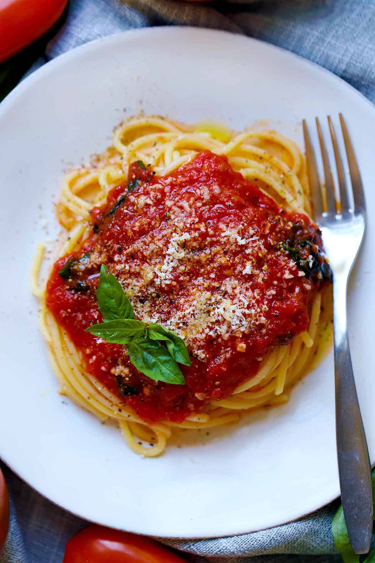 Overhead photo of a plate with spaghetti and tomato sauce topped with a basil leaf and parmesan cheese and a fork next to it.