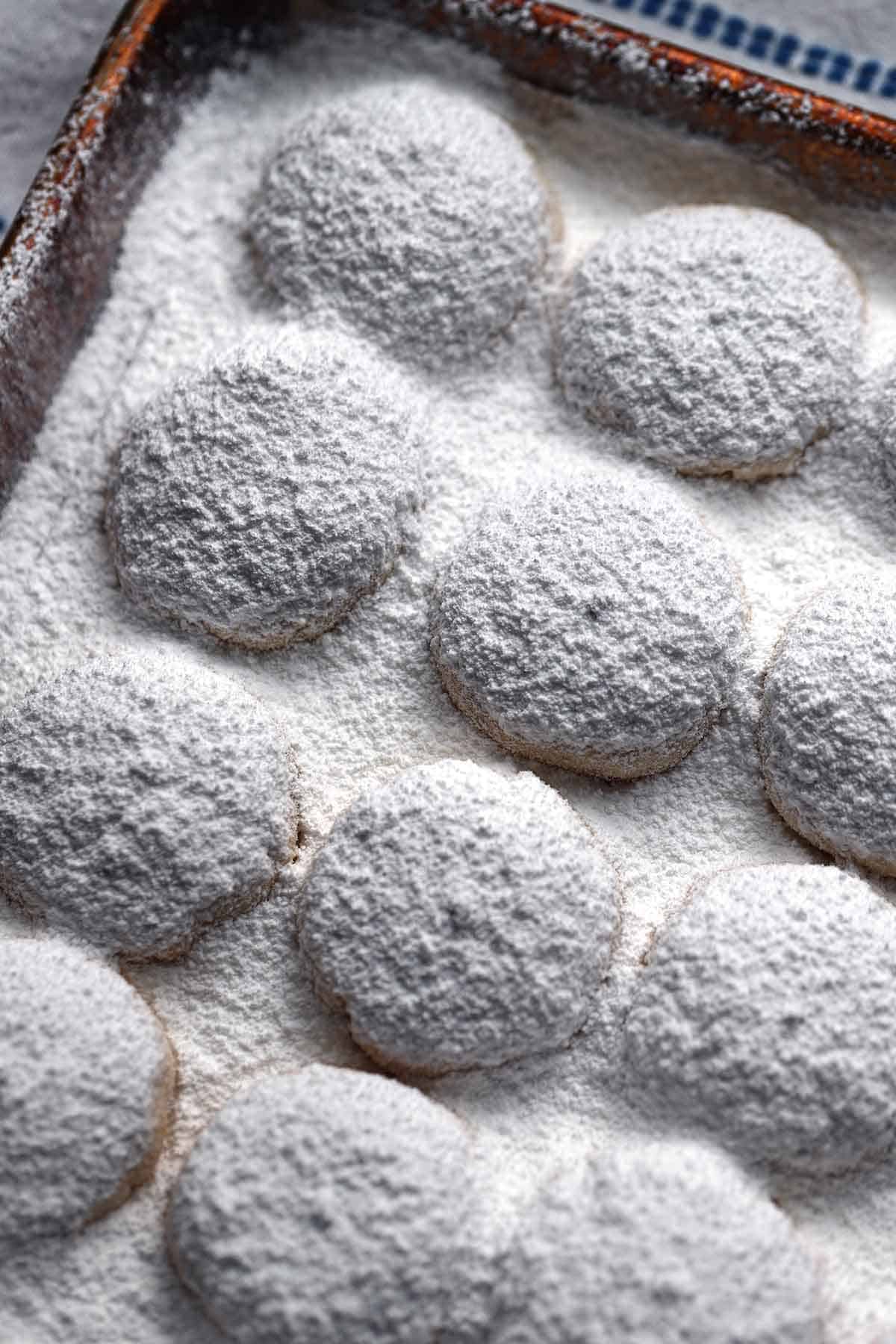 Kourambiedes on a cookie sheet coated in powdered sugar, cooling after baking.