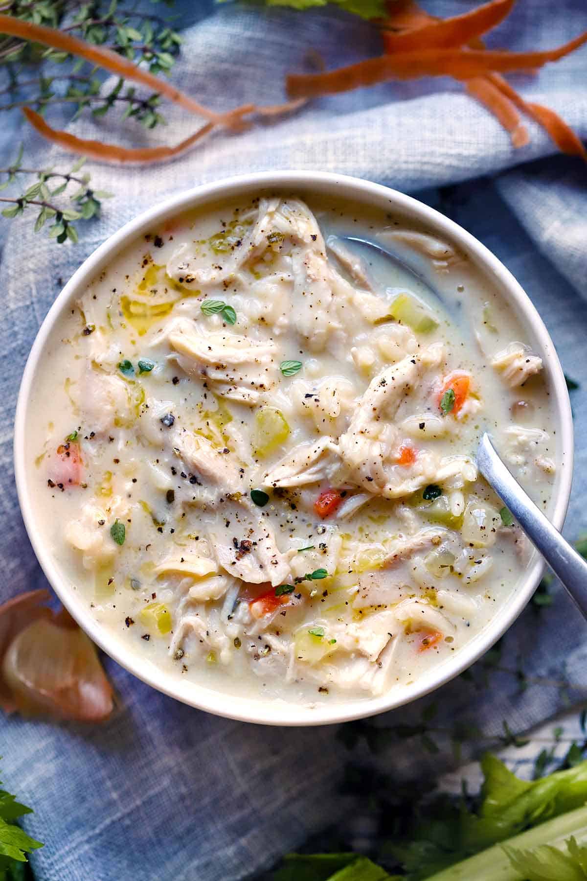 Overhead photo of a bowl of creamy chicken and rice soup with a spoon in it and carrots and celery and thyme scattered around.
