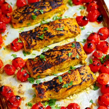 Square photo of tandoori salmon filets on a sheet pan with burst cherry tomatoes.