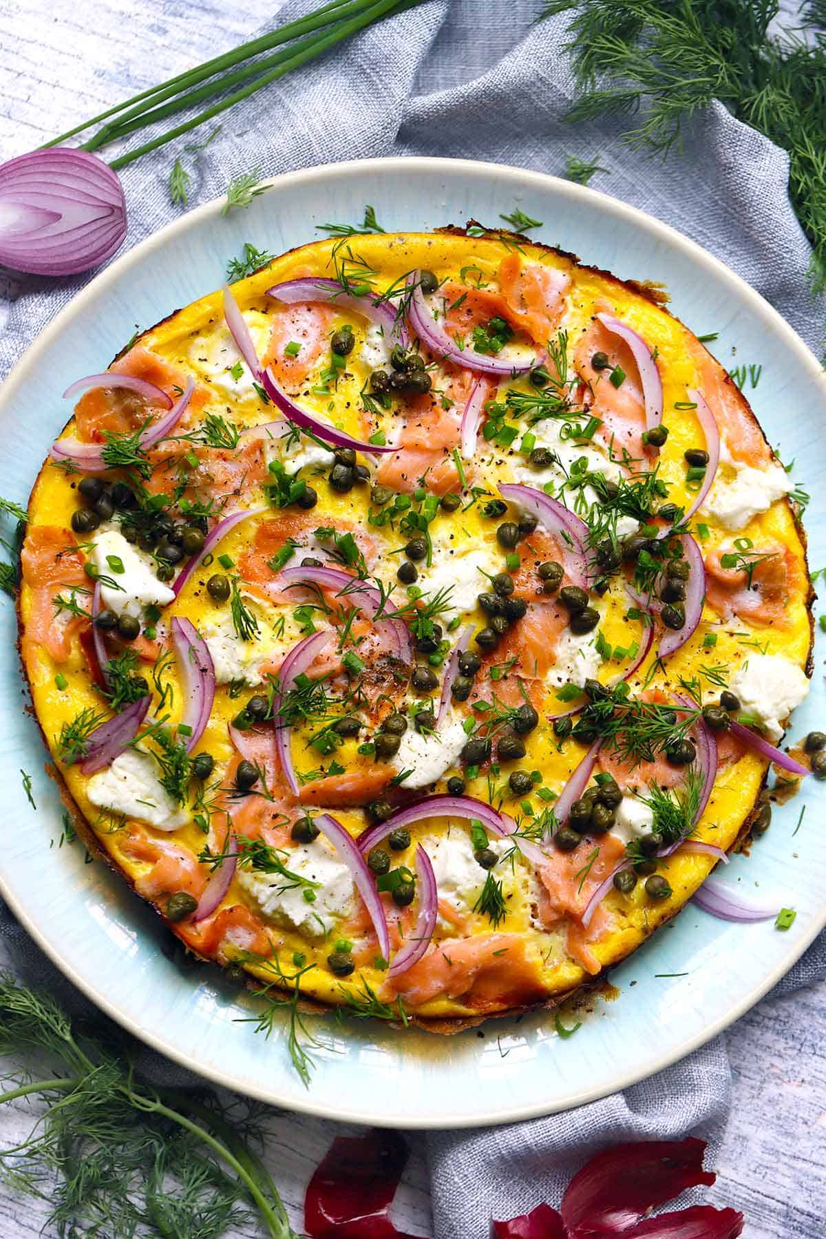 Overhead view of an open faced omelette or stovetop frittata topped with smoked salmon, cream cheese, capers, re onions, and herbs, with dill and red onion scattered around.