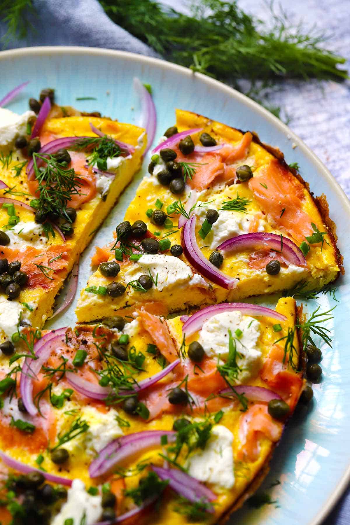An open faced omelette topped with smoked salmon and cream cheese with a slice cut out on a blue plate.
