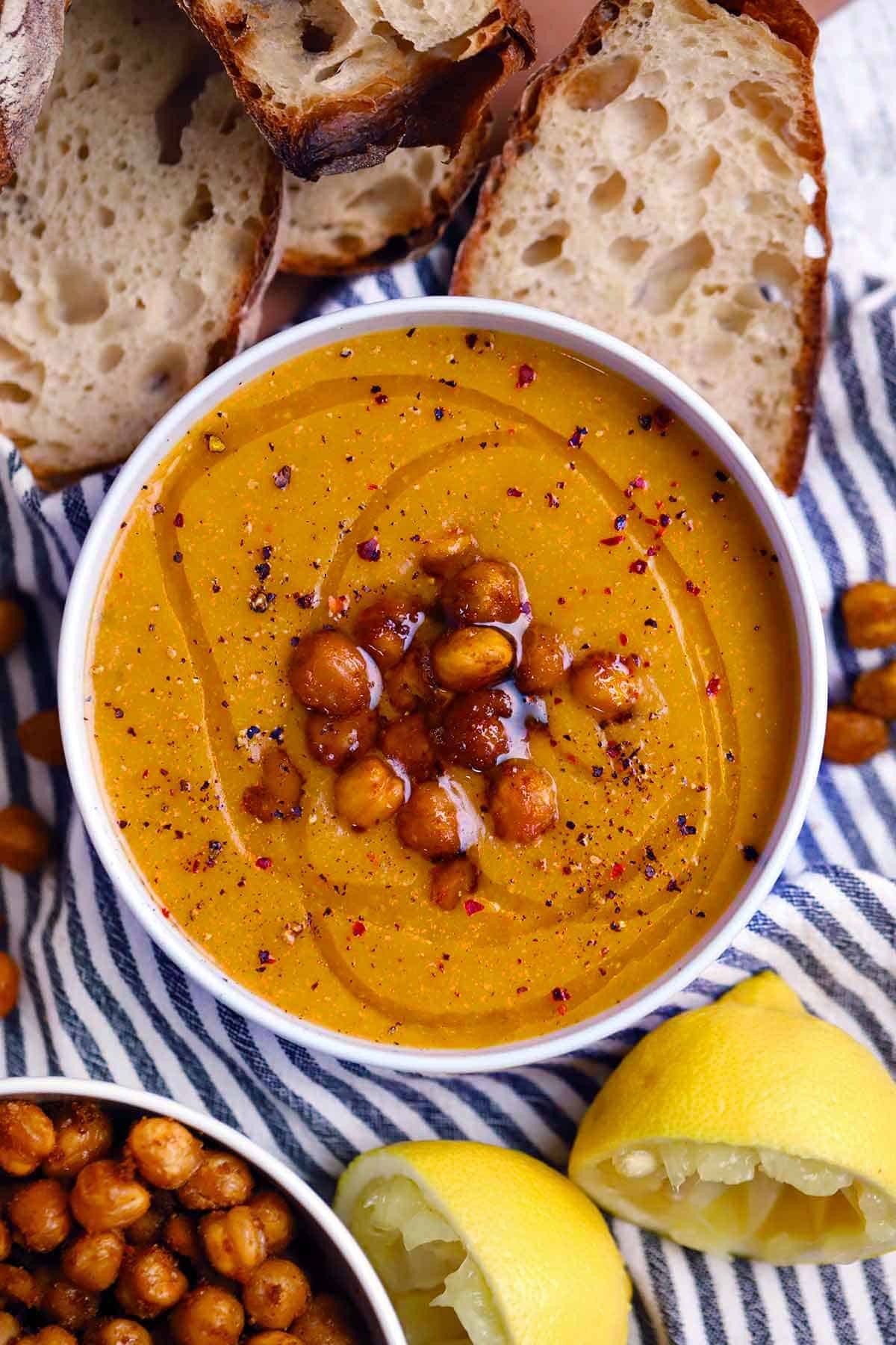 Overhead view of a bowl of chickpea soup drizzled with olive oil and topped with roasted chickpeas, with sliced bread, lemon halves, and a bowl of roasted chickpeas scattered around on a blue and white striped towel.