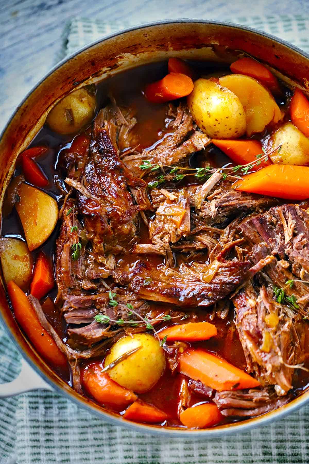 Pot roast in a Dutch Oven with carrots and potatoes, garnished with thyme sprigs, with the meat shredded and added to the liquid in the pot.