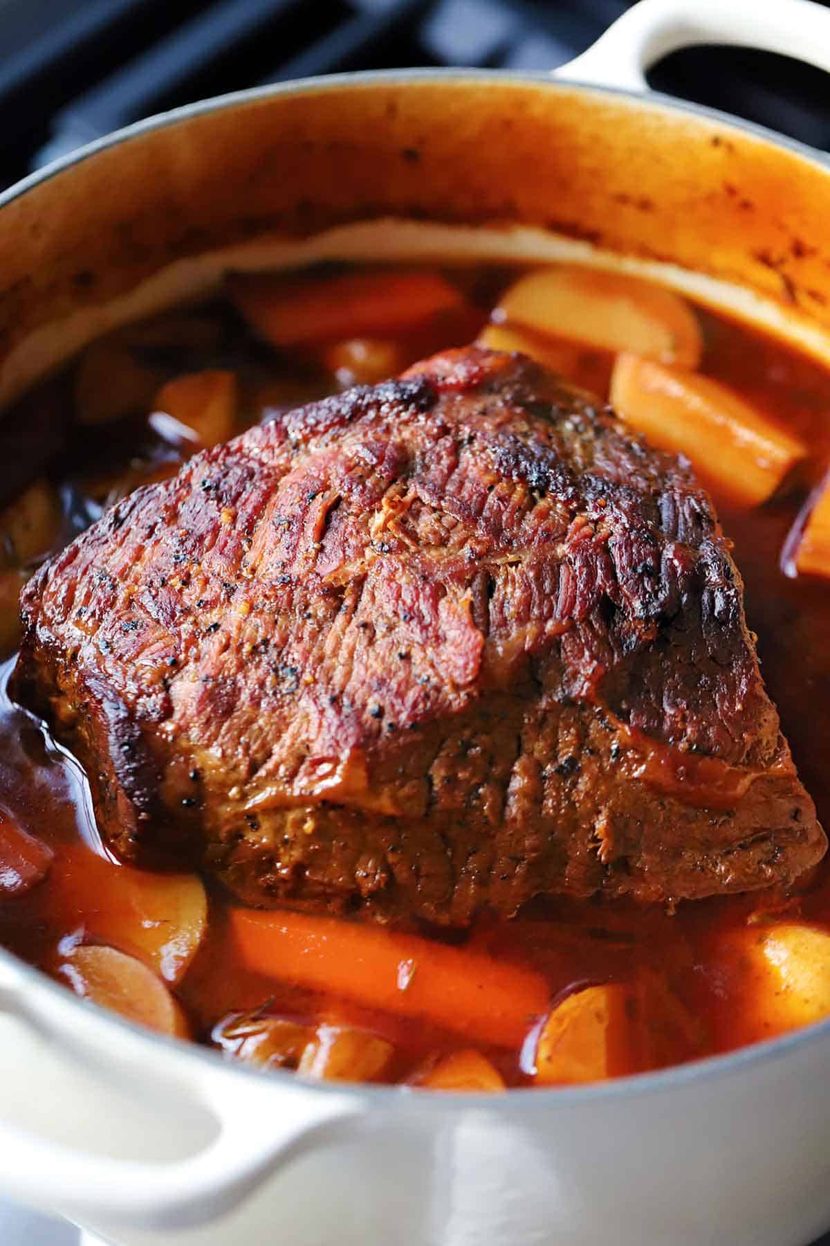 A bottom round pot roast cooked in a dutch oven, with carrots and potatoes, still whole and not shredded yet.