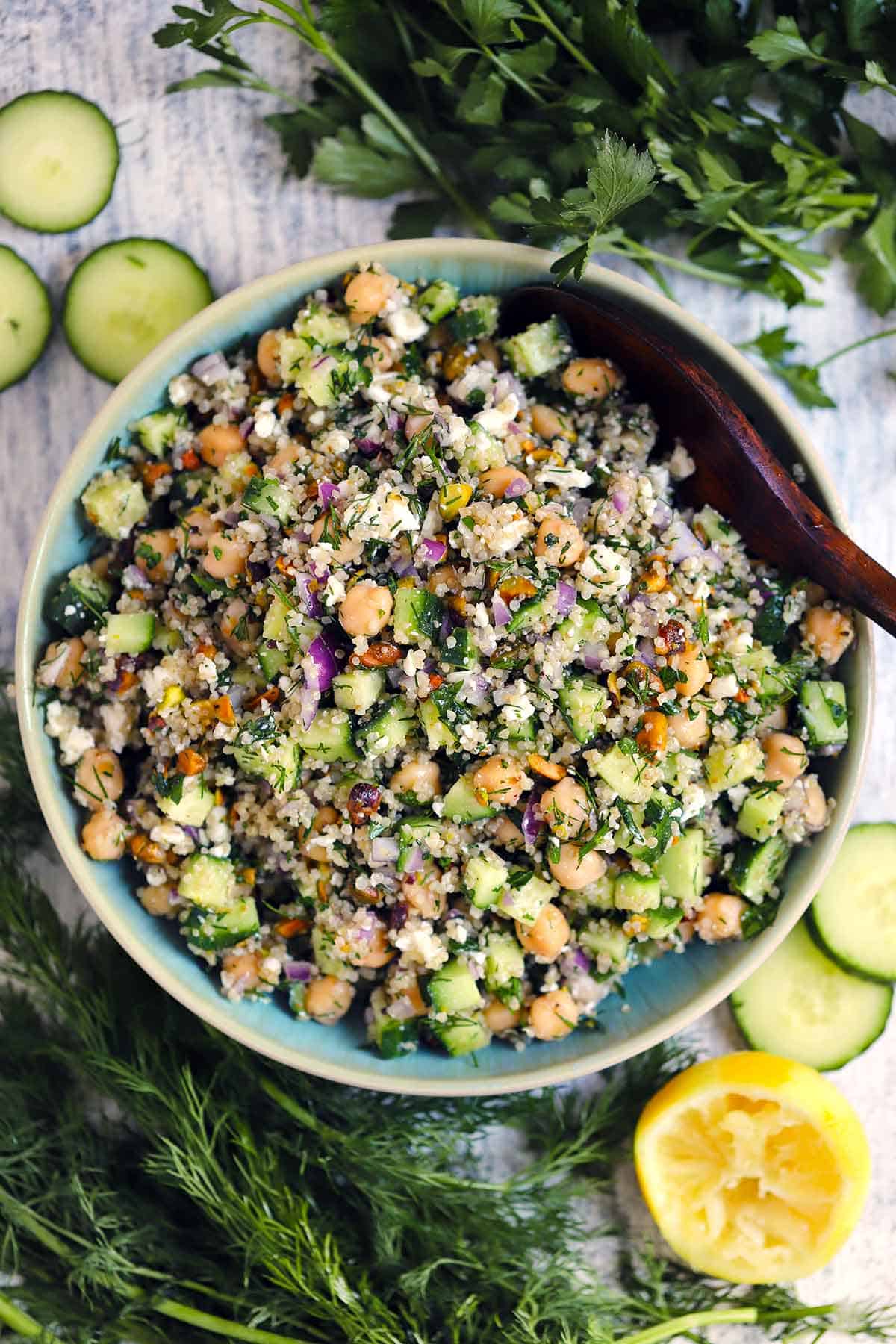 Overhead photo of a bowl of Jennifer Aniston Salad with a wooden spoon, with cucumbers, lemon, and herbs scattered around.