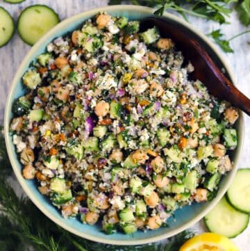 Square photo of an overhead shot of the Jennifer Aniston Salad in a blue bowl with a wooden spoon in it and ingredients scattered around.
