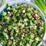 Square cropped photo of a close up overhead view of cold farro salad with cucumbers, feta cheese, herbs, capers, and green onions, with a spoon nestled in.