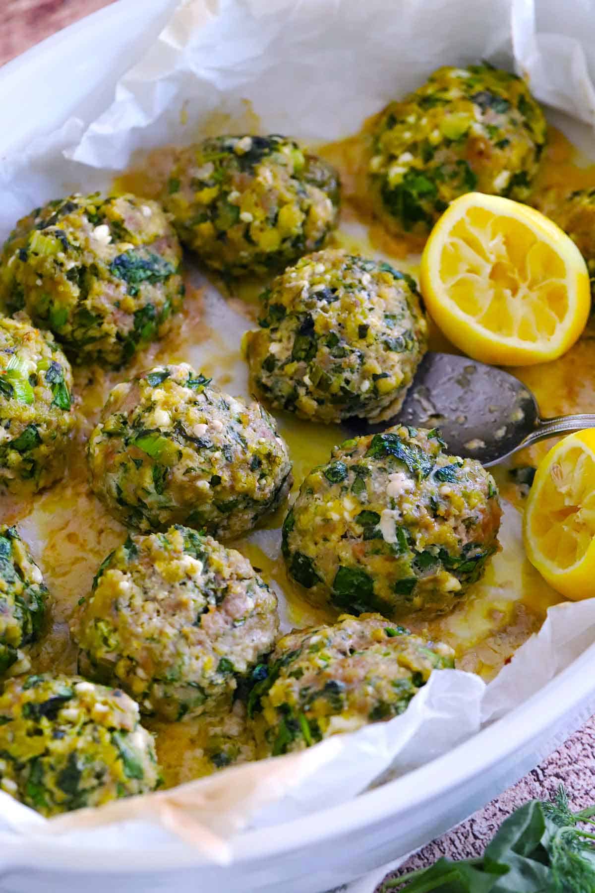 Baked spanakopita meatballs in a baking dish on parchment paper with lemon halves and a spoon scooping one out.