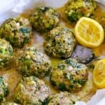 Square photo of spanakopita meatballs in a baking dish on parchment paper, with lemon halves, and a spoon lifting a meatball out.