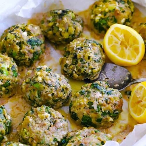Square photo of spanakopita meatballs in a baking dish on parchment paper, with lemon halves, and a spoon lifting a meatball out.