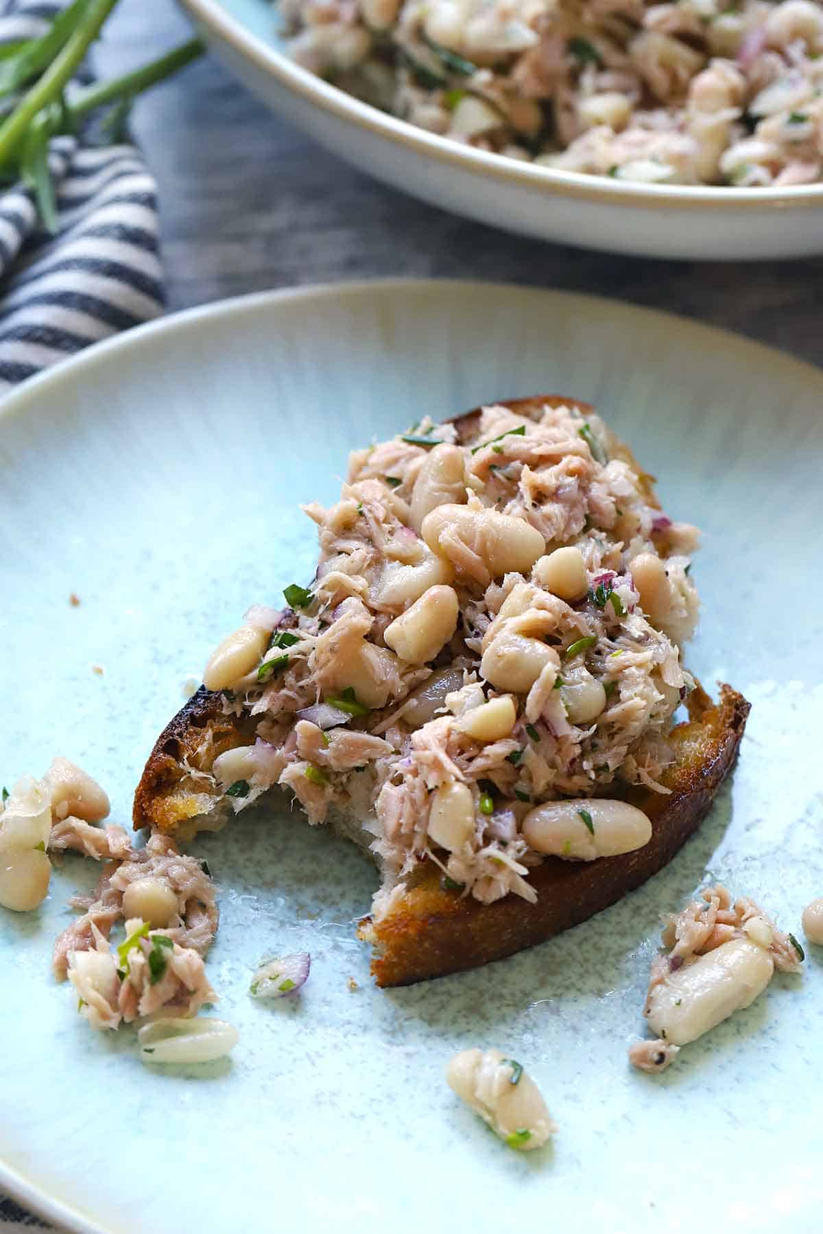 A piece of toasted bread with white bean and tuna salad on top for an open faced sandwich on a blue plate.