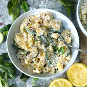 Square photo of a close up overhead photo of a bowl of lemon ricotta pasta with zucchini and basil with ingredients scattered around in a white bowl.