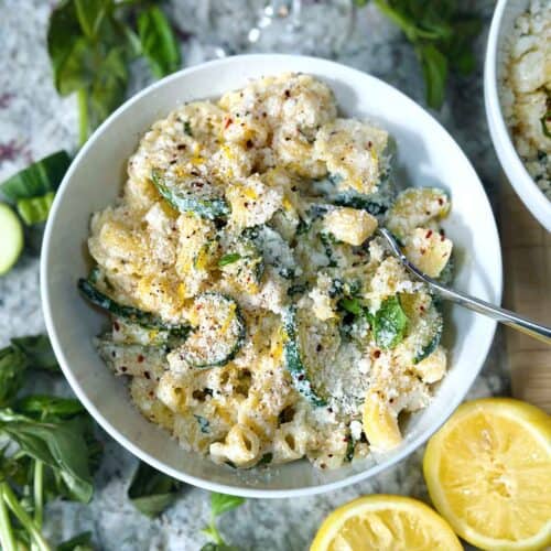 Square photo of a close up overhead photo of a bowl of lemon ricotta pasta with zucchini and basil with ingredients scattered around in a white bowl.