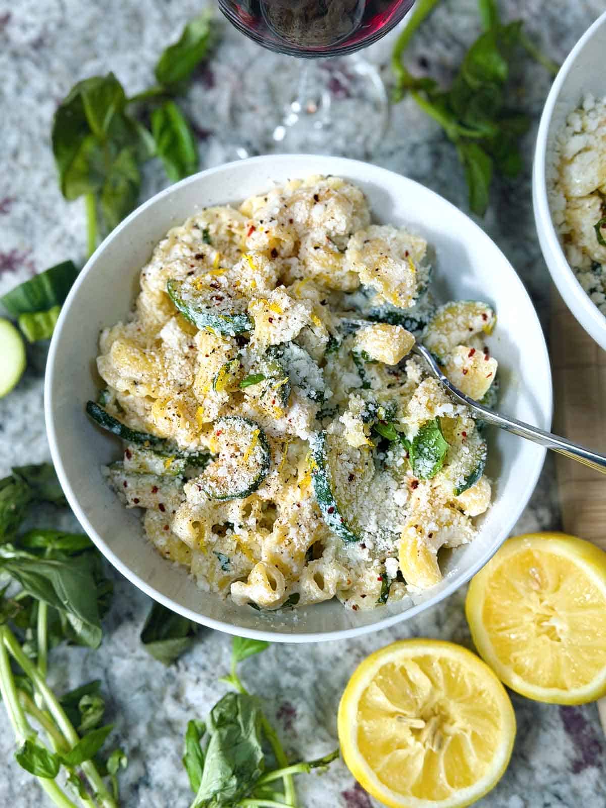 Overhead photo of a bowl of lemon ricotta pasta with zucchini and basil with ingredients scattered around in a white bowl.