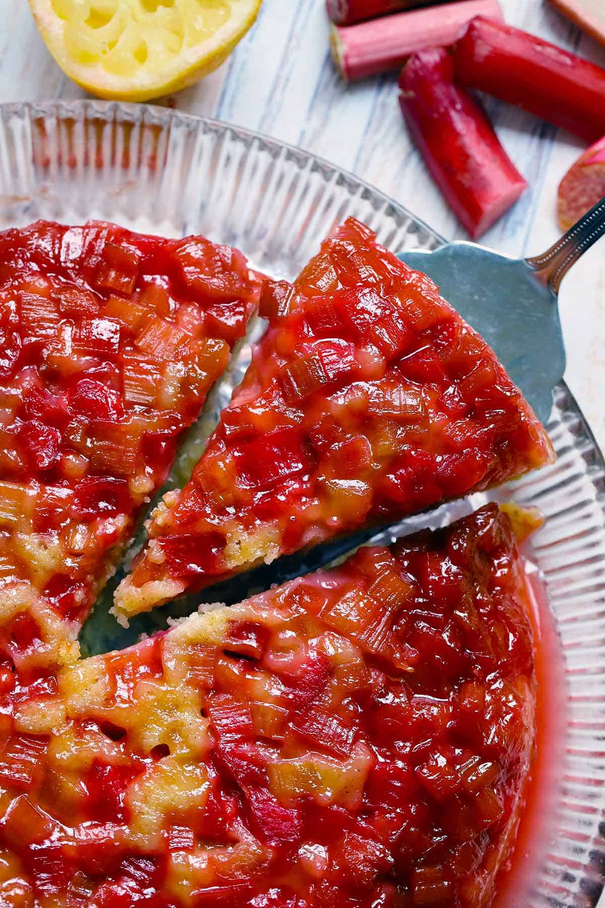 A slice of upside down rhubarb cake being taken out of the circular whole cake, which is on a glass platter, with rhubarb and lemon scraps scattered around.