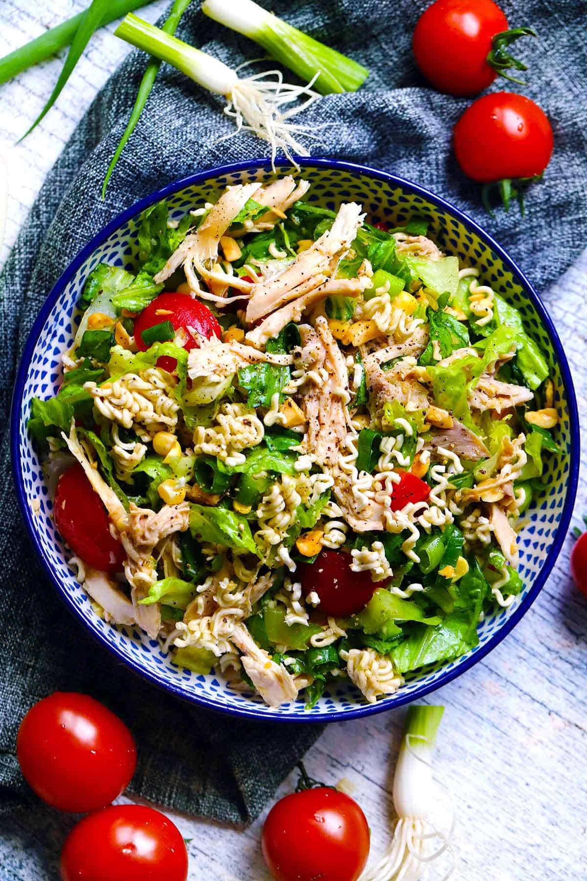 Overhead photo of a bowl of crunchy ramen noodle salad, with tomatoes and green onions scattered around, in a blue patterned bowl.